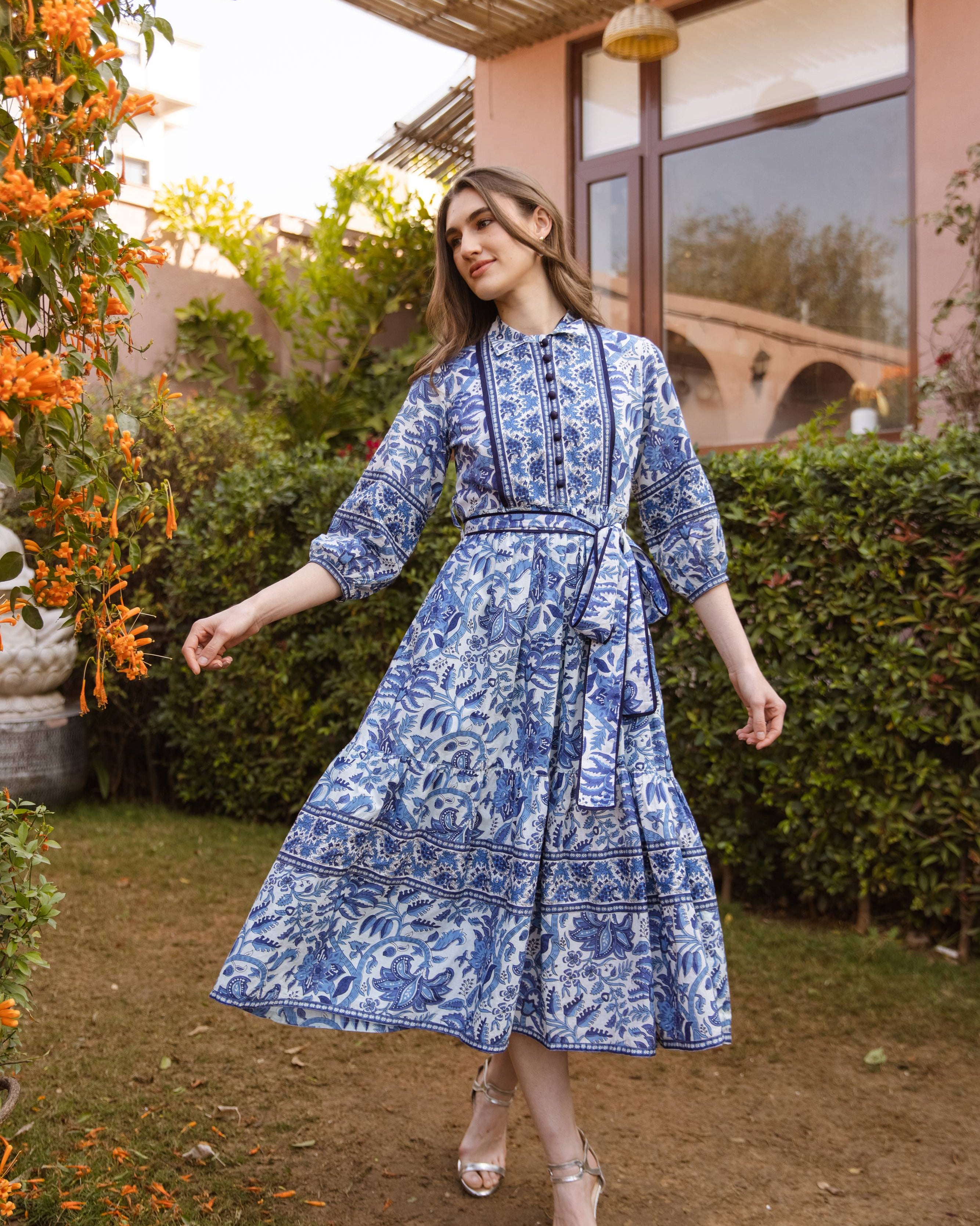 Woman in a blue patterned dress standing outdoors near a building with greenery.