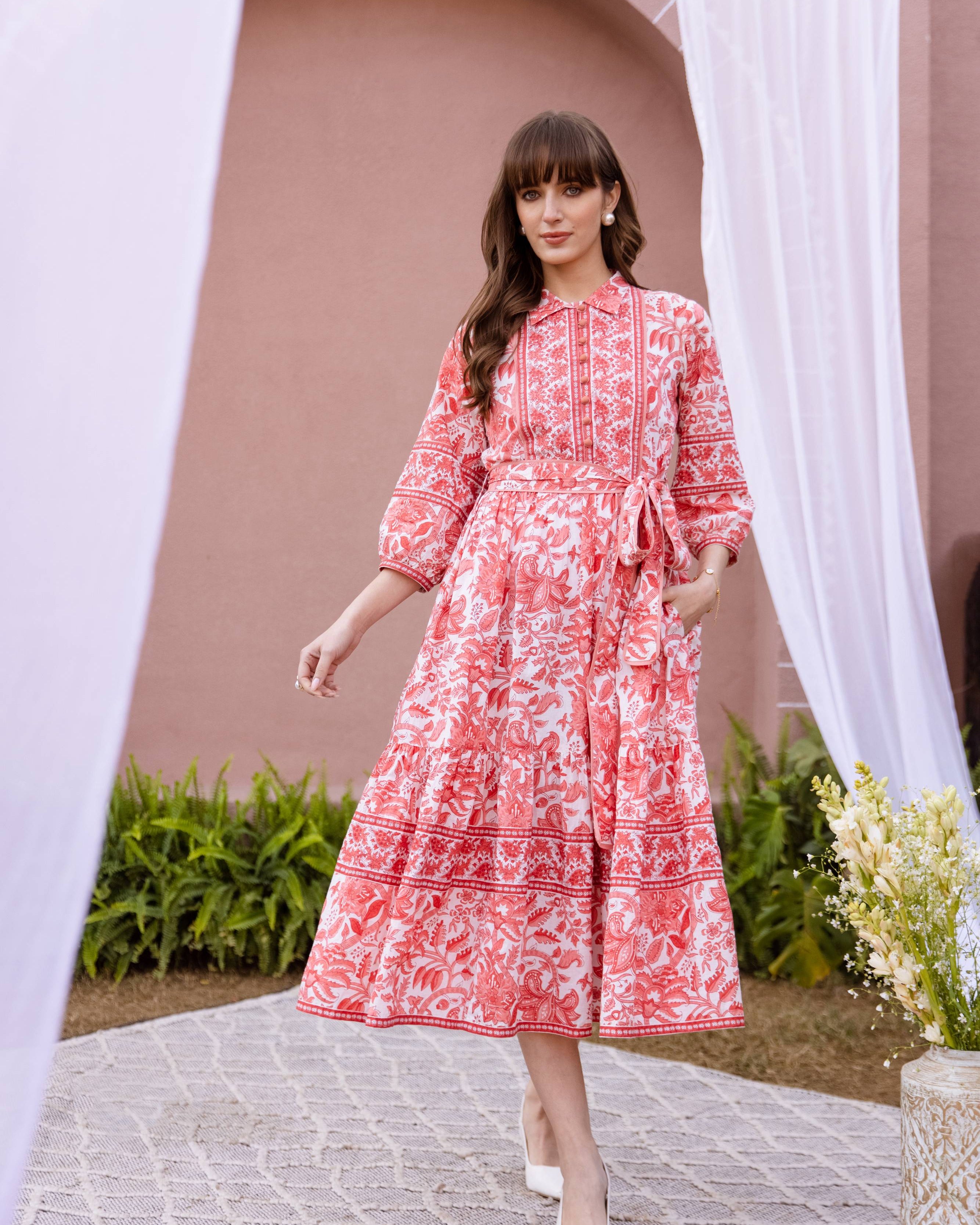 Woman in a orange and white patterned dress standing outdoors with a decorative arch and plants in the background.