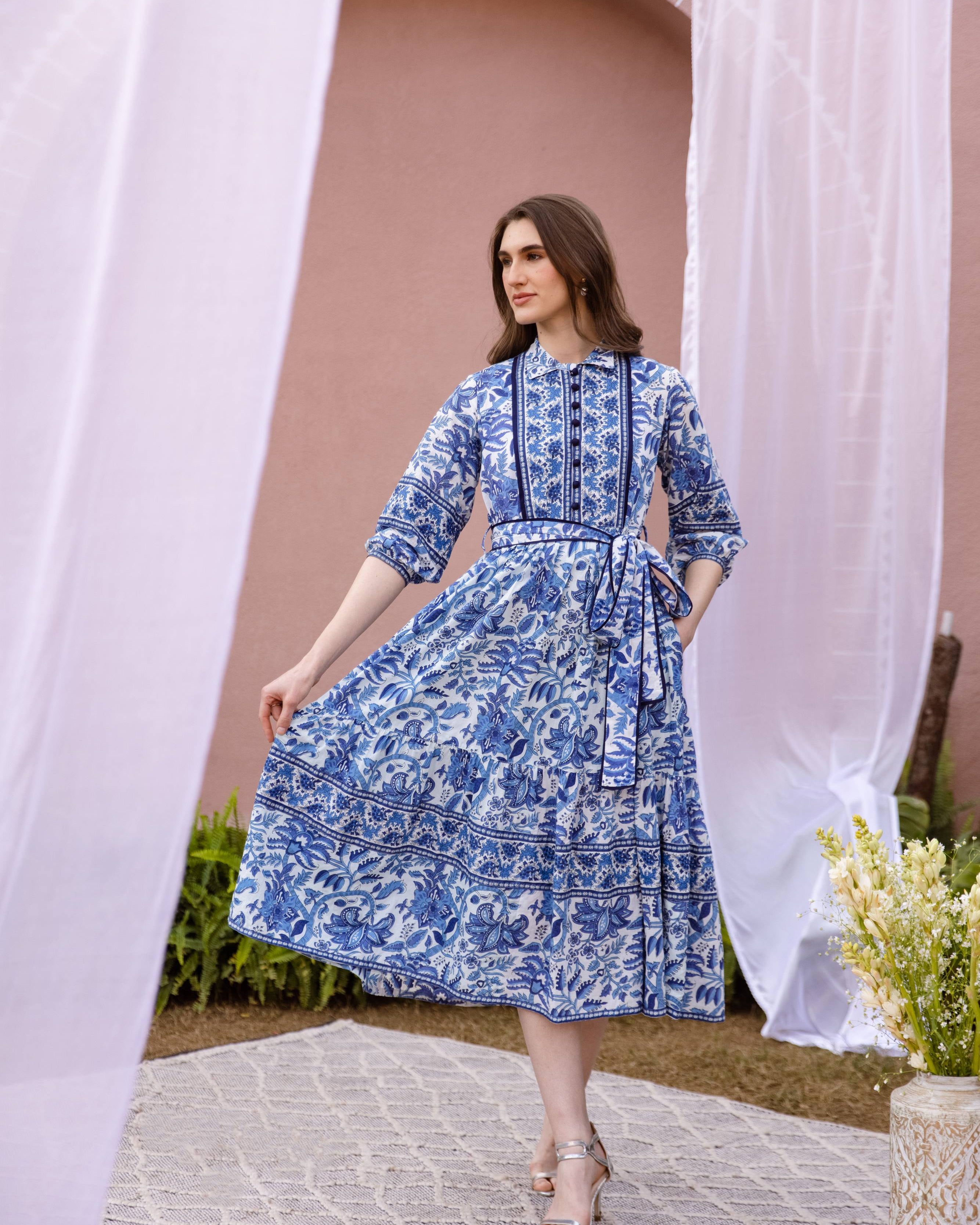 Woman in a blue floral dress standing in front of a pink wall with white curtains.