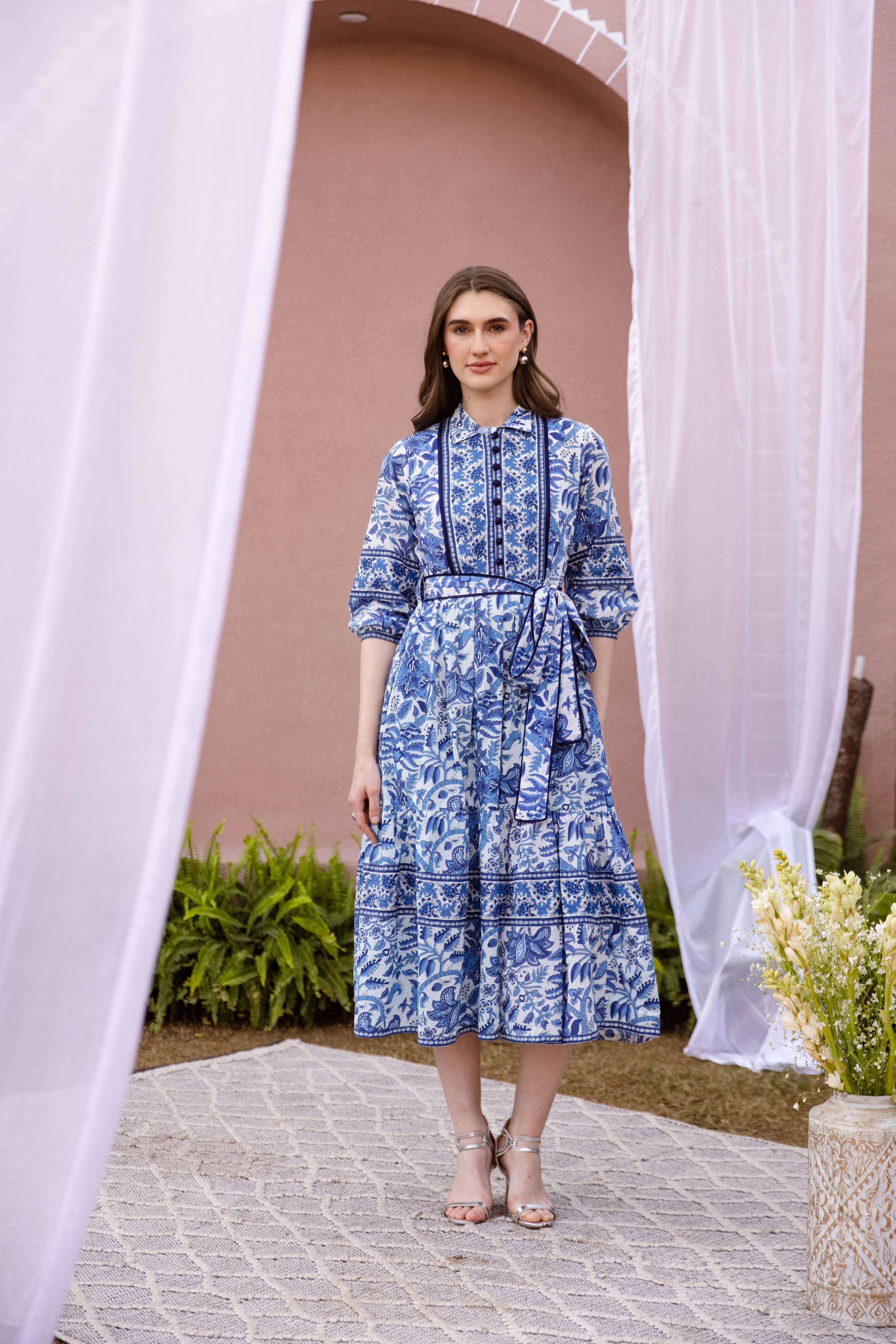 Woman in a blue floral dress standing under a white archway with a pink wall and greenery in the background.