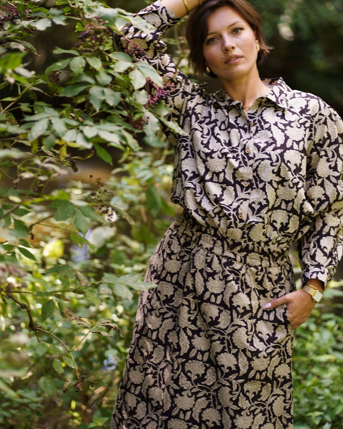 Woman wearing a patterned shirt and skirt standing among green foliage