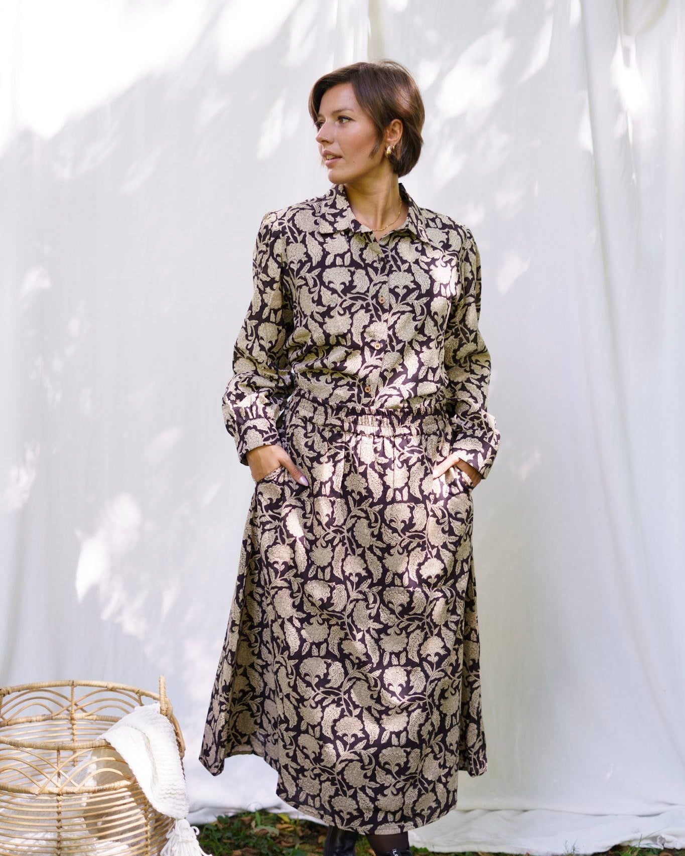 Woman wearing a long, patterned dress standing against a white curtain.