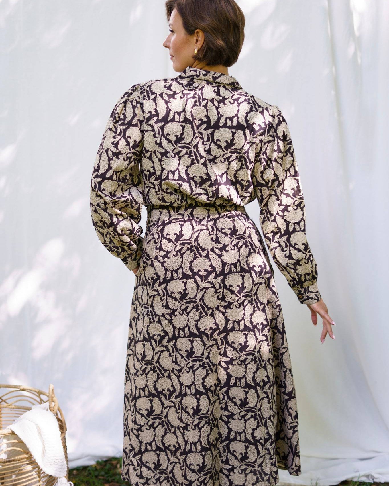 Woman wearing a long, patterned dress standing against a white curtain.