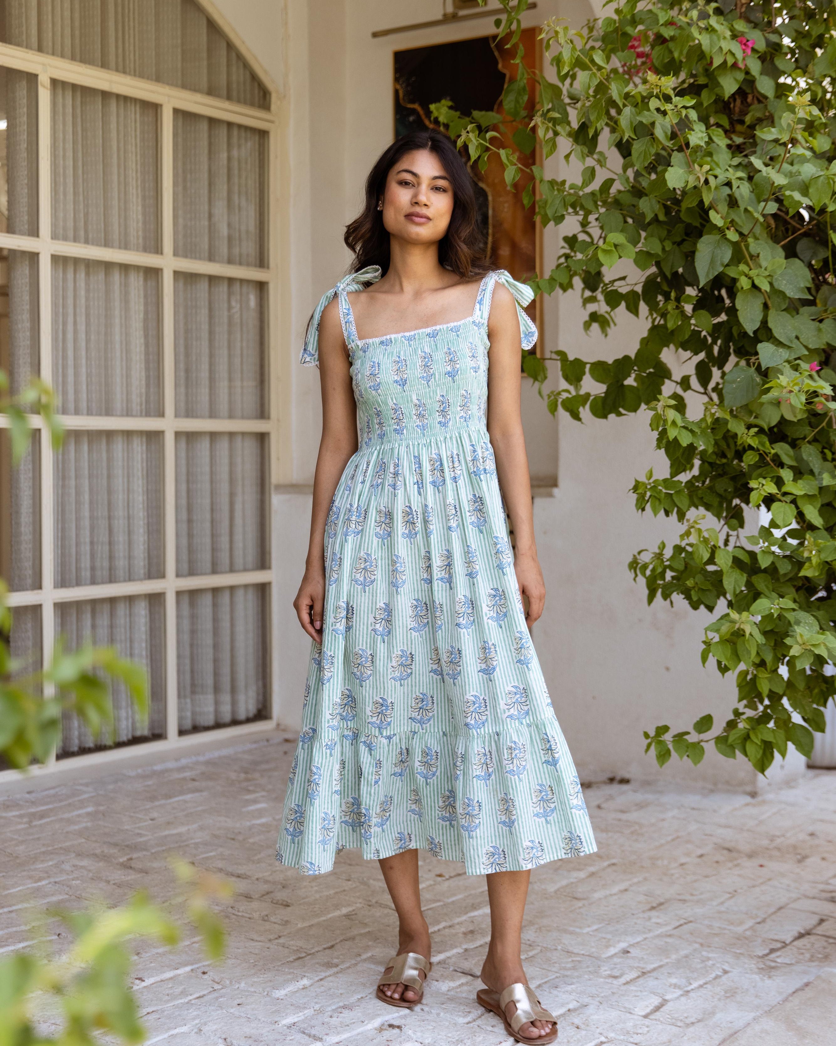A woman standing in an indoor setting wearing a mint green and blue floral dress with tie-up straps and lace details.
