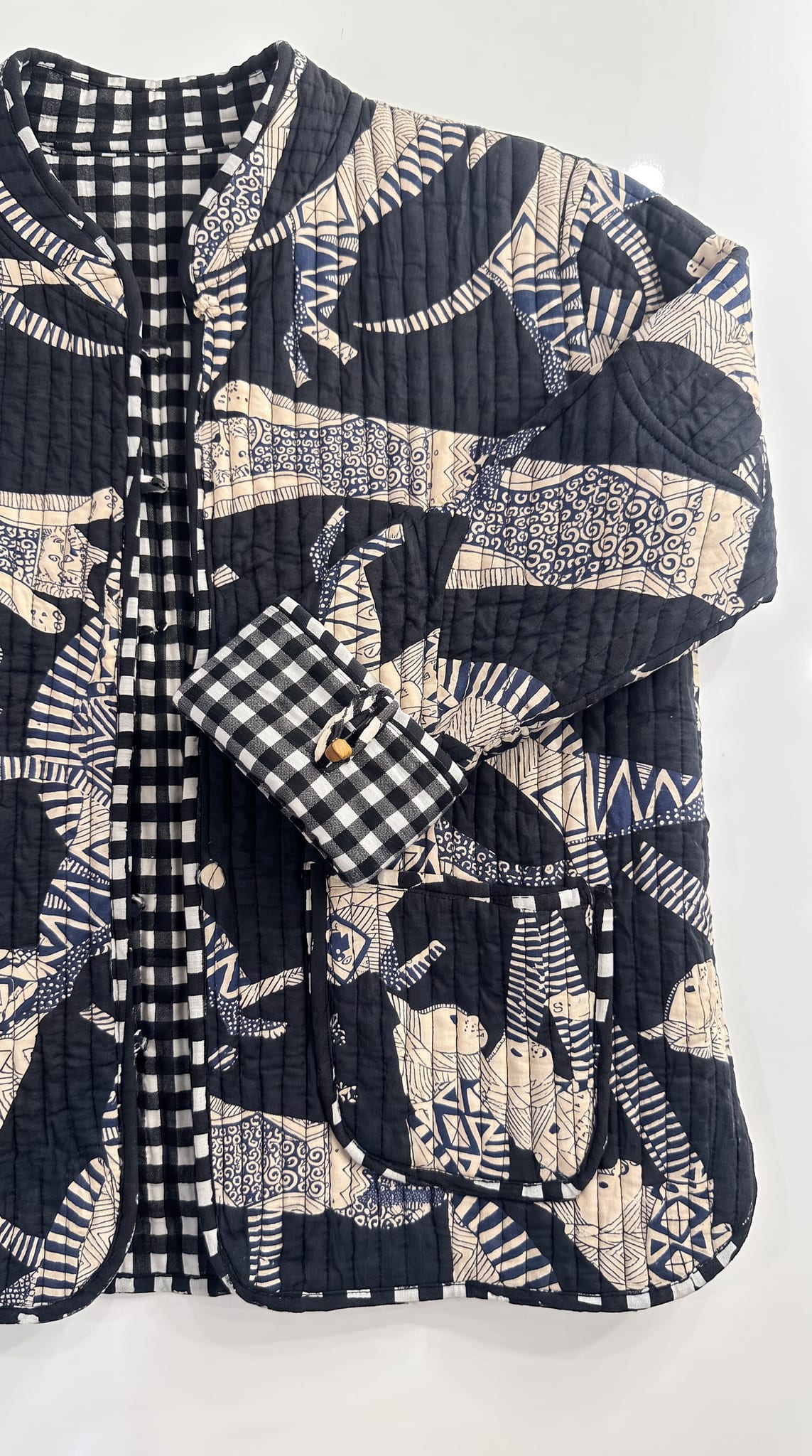 Close-up flat lay of Blue Cat Chaos Jacket showing quilted cotton texture, navy and cream cat print, gingham cuff, wooden toggle button, and patch pocket detail