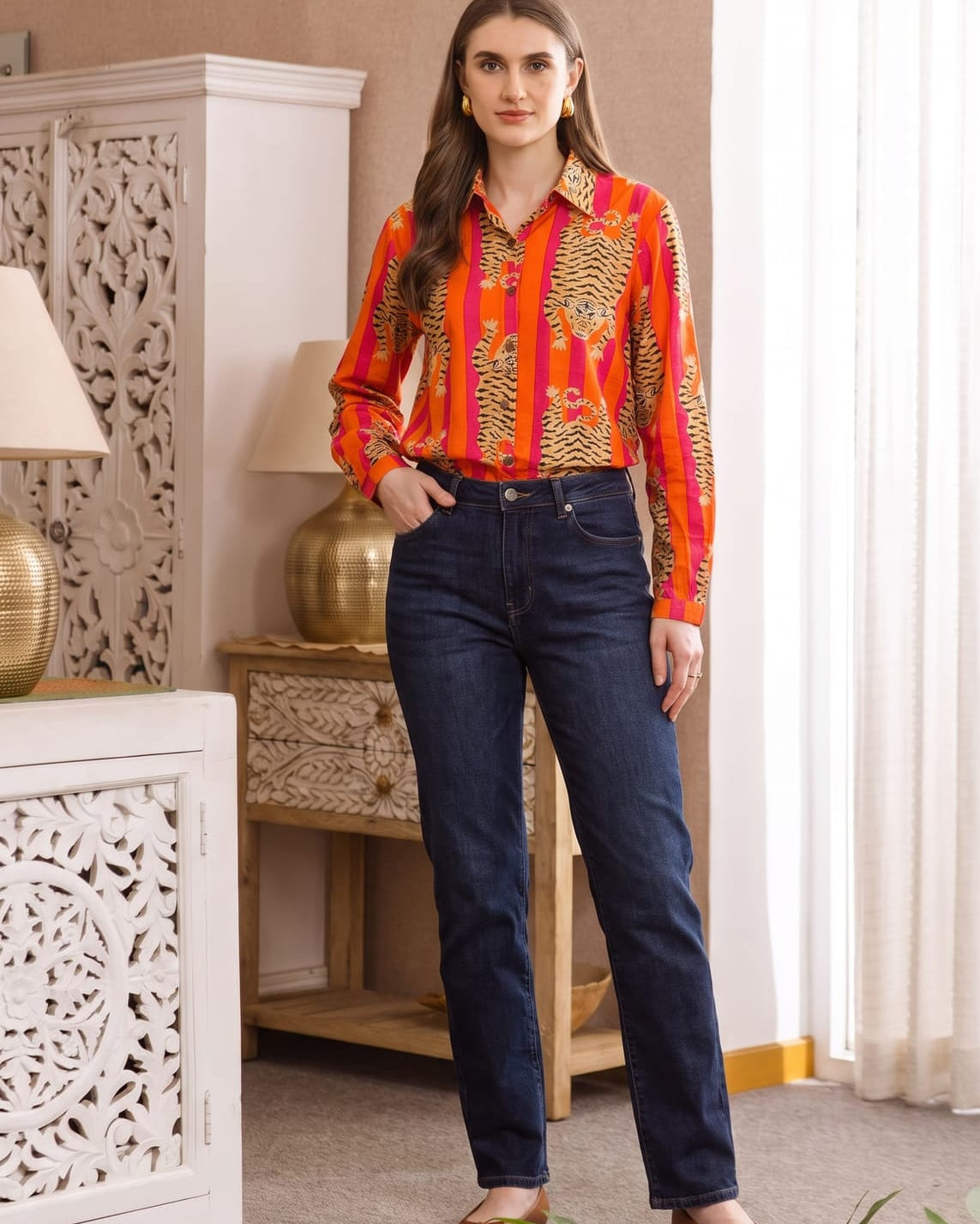 Full-length front view of model wearing the Tibetan Tiger Blouse, an orange and pink striped tiger-print cotton blouse, tucked into dark denim jeans with tan flats, in a home interior with carved wood furniture