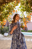 Woman in a blue patterned dress standing under a tree with orange flowers.