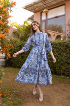 Woman in a blue patterned dress standing outdoors near a building with greenery.