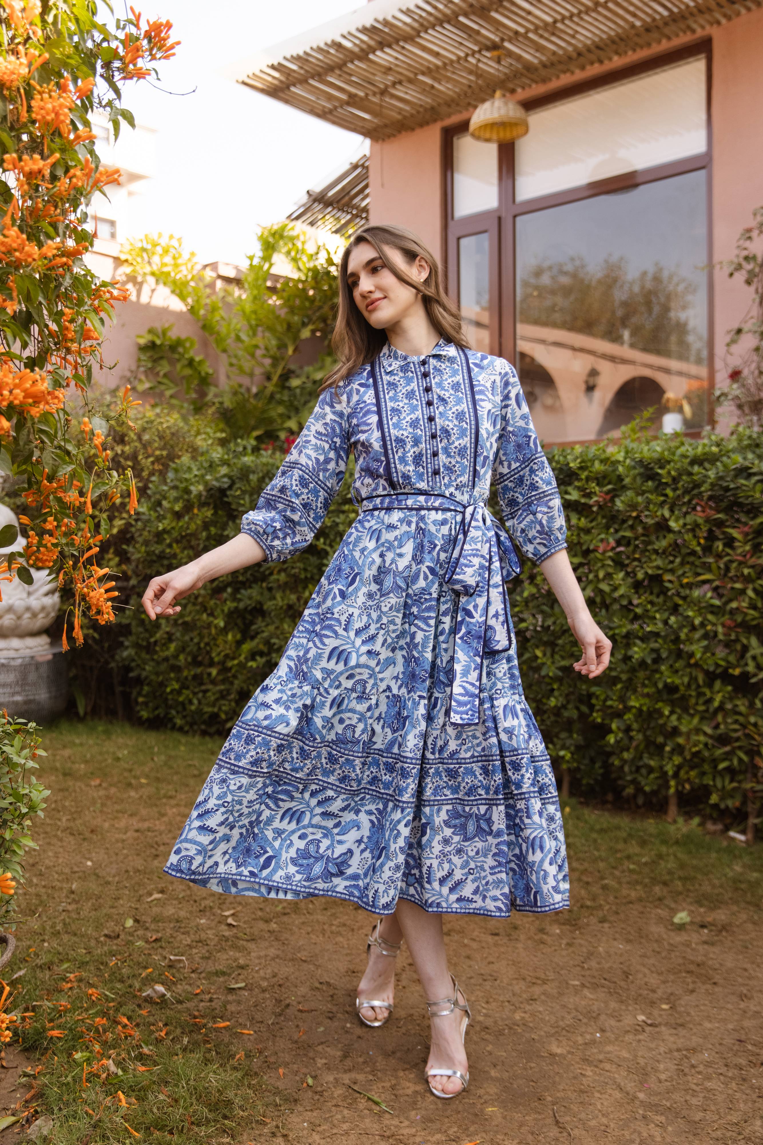 Woman in a blue patterned dress standing outdoors near a building with greenery.