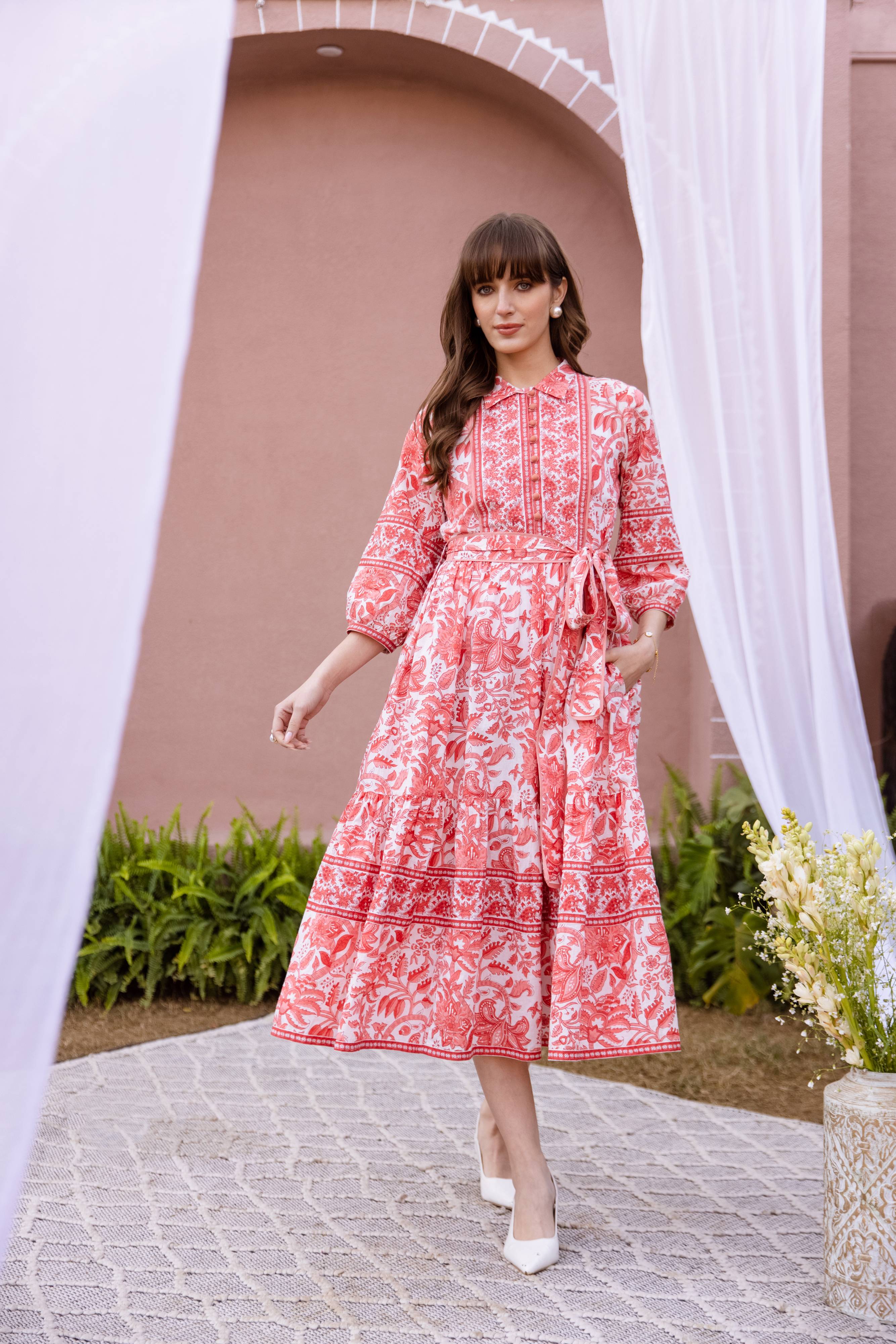 Woman in a orange and white patterned dress standing outdoors with a decorative arch and plants in the background.