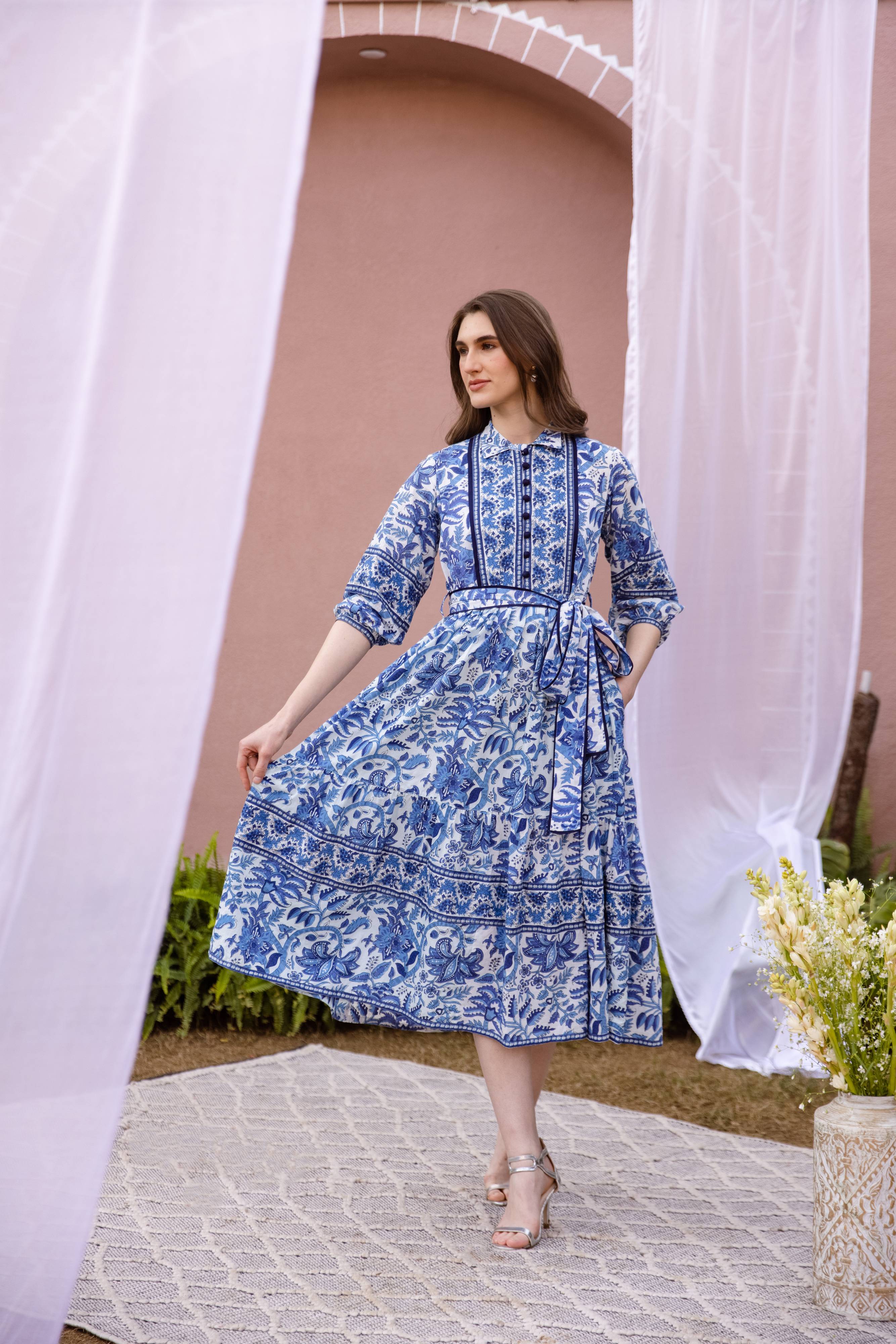 Woman in a blue floral dress standing in front of a pink wall with white curtains.