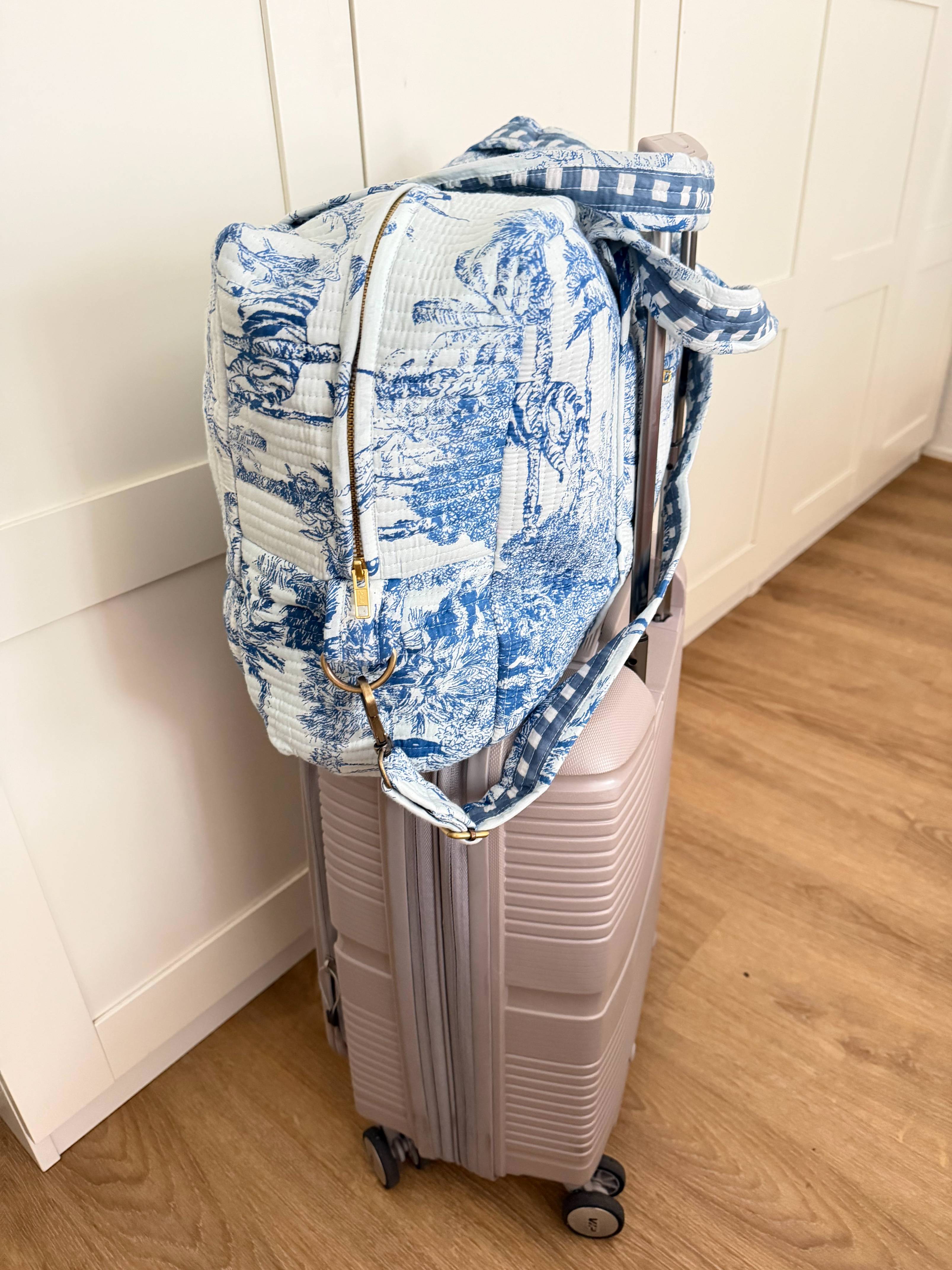 Blue and white toile quilted duffel bag with gingham-lined strap secured on top of a mauve hardshell suitcase in a hallway, shown from the side with gold zipper and clasp visible