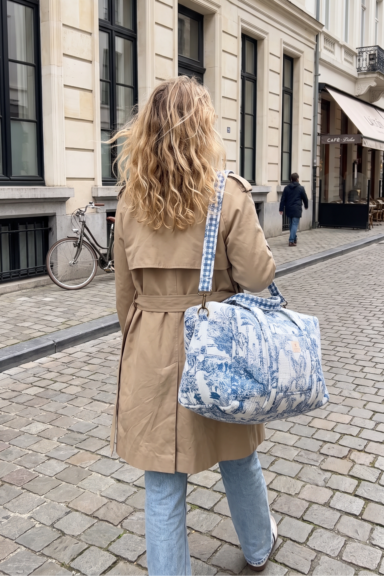 Woman in a beige trench coat and jeans walking down a cobblestone European street, carrying the blue and white toile duffel bag on her shoulder by its blue gingham crossbody strap