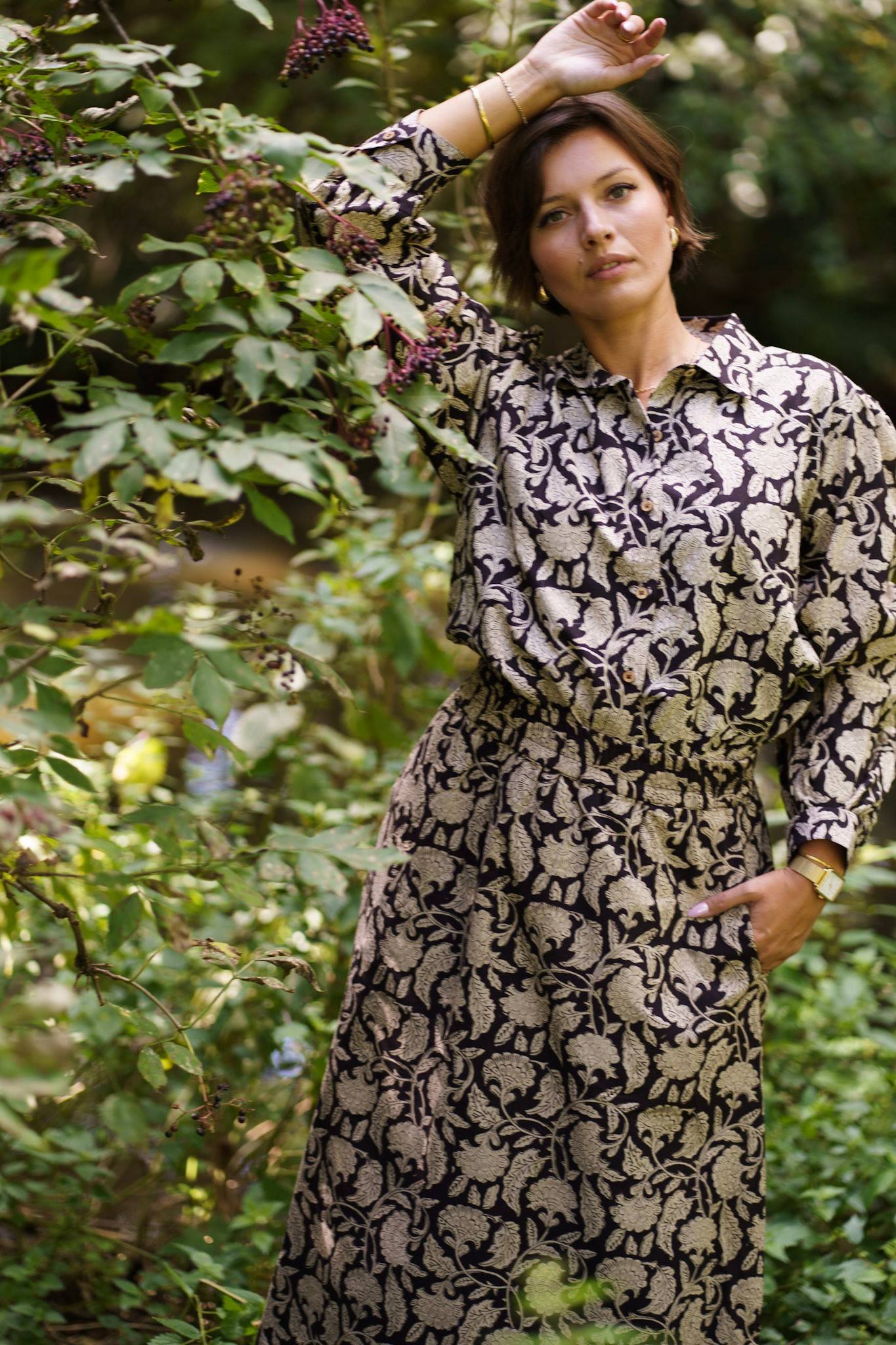 Woman wearing a patterned shirt and skirt standing among green foliage
