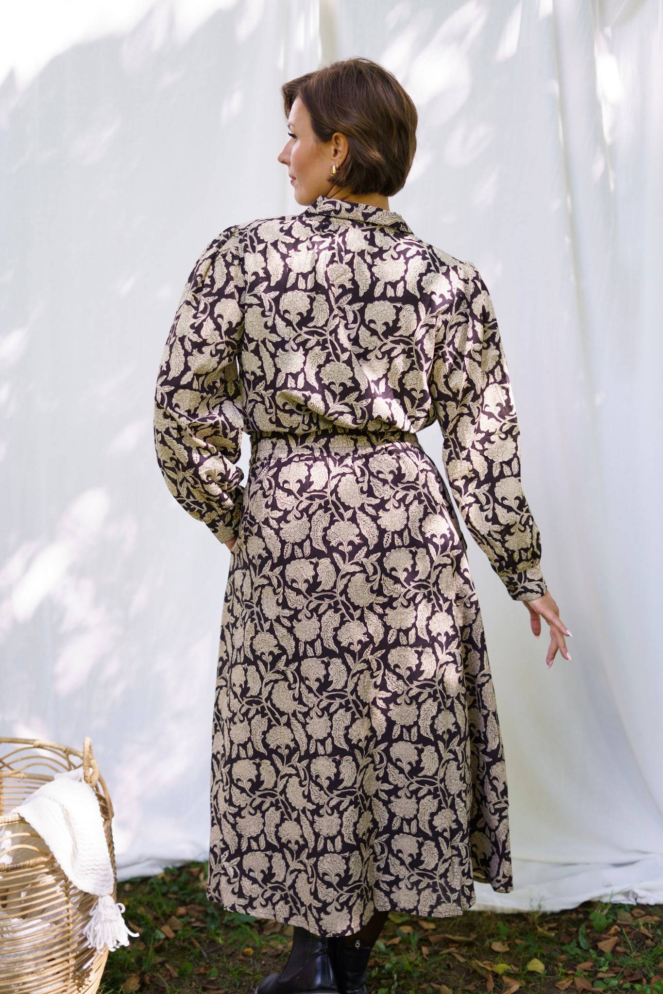 Woman wearing a long, patterned dress standing against a white curtain.