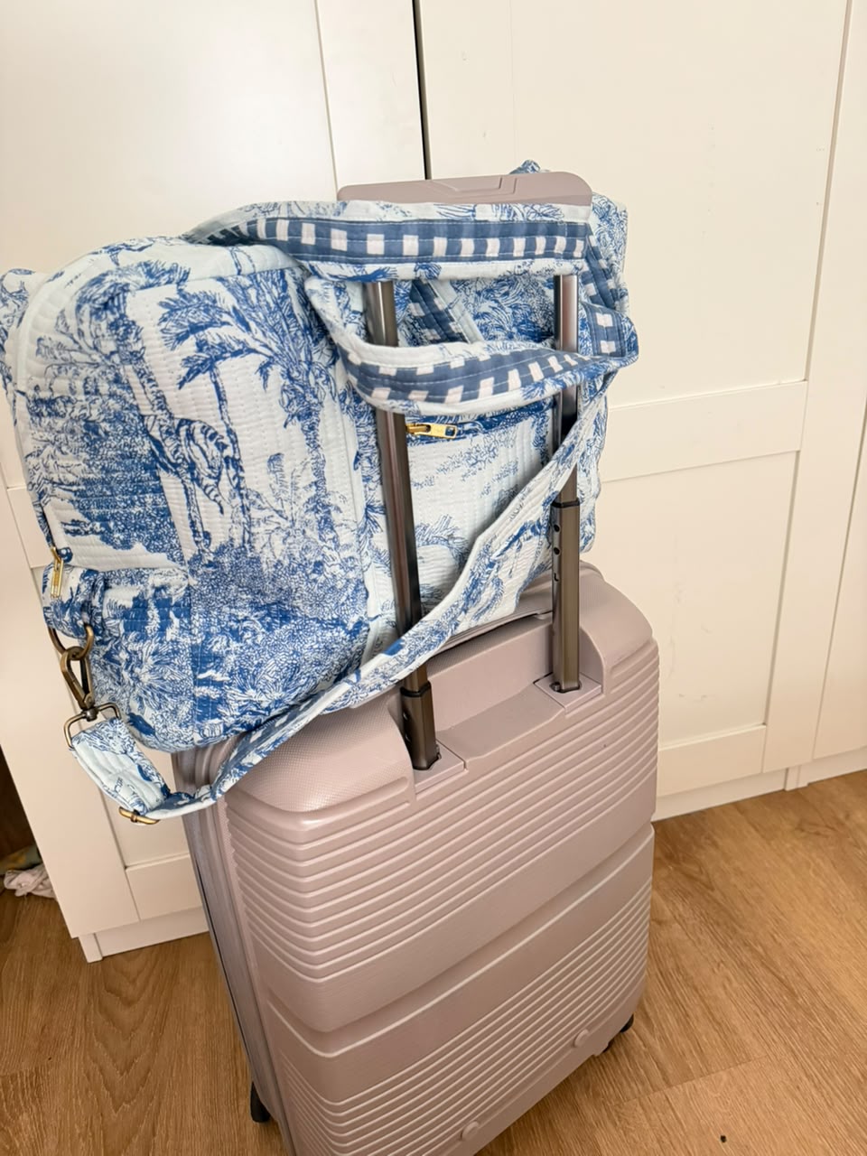 A blue and white toile de Jouy patterned duffel bag with blue-and-white gingham trim, draped over a light pink hard-shell suitcase in an indoor setting.