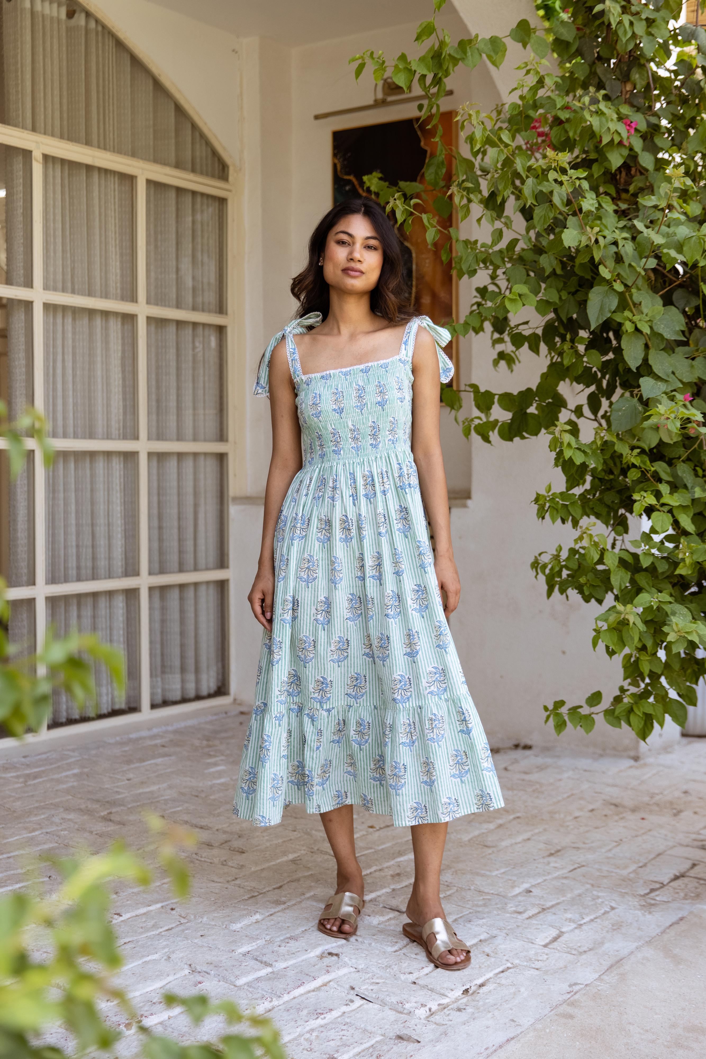 A woman standing in an indoor setting wearing a mint green and blue floral dress with tie-up straps and lace details.