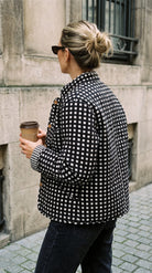 Reversible side of the Blue Cat Chaos Jacket in black and white gingham check, back view worn with wooden toggle buttons visible, model holding a coffee cup