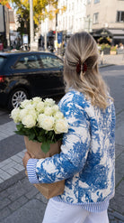 Blue Jardin Jacket in blue and white toile print, back view, worn with white jeans while carrying a bouquet of white roses on a city street
