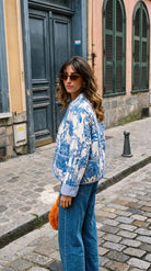 Side/back view of the Blue Jardin Jacket in blue and white toile print, worn with blue jeans and an orange crochet bag on a cobblestone street