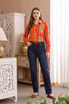 Full-length front view of model wearing the Tibetan Tiger Blouse, an orange and pink striped tiger-print cotton blouse, tucked into dark denim jeans with tan flats, in a home interior with carved wood furniture